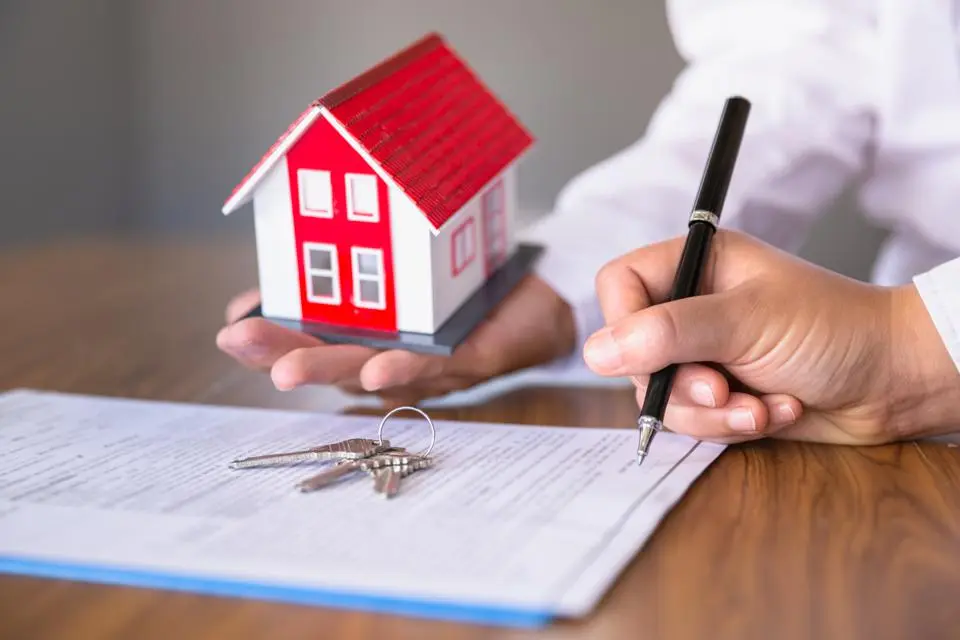 A hand holding a house and the other hand signing in a document with keys on it